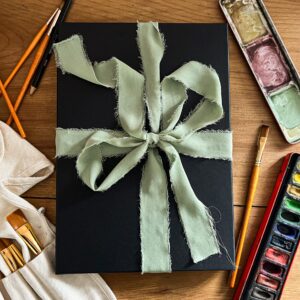Print and Bookmark Gift Bundle image of gift box wrapped in sage green ribbon and surrounded by watercolour paints and brushes