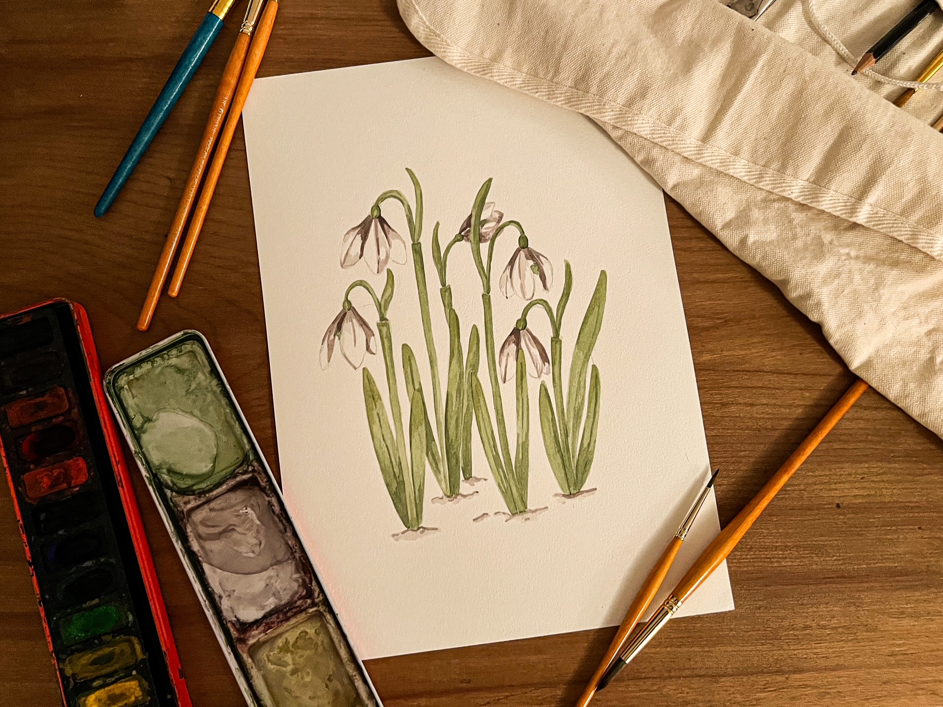 Image from above of Snowdrop painting by Emma Macdonald, artist behind Pippin Prints, shown on desk surrounded by paint brushes and paints