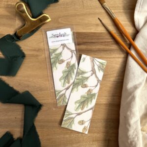 Photograph of Acorns Bookmark by Pippin Prints shown both with and without packaging and surrounded by a dark green ribbon and paint brushes.