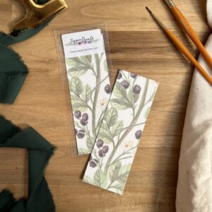 Photograph of Brambles Bookmark by Pippin Prints shown both with and without packaging and surrounded by a dark green ribbon and paint brushes.