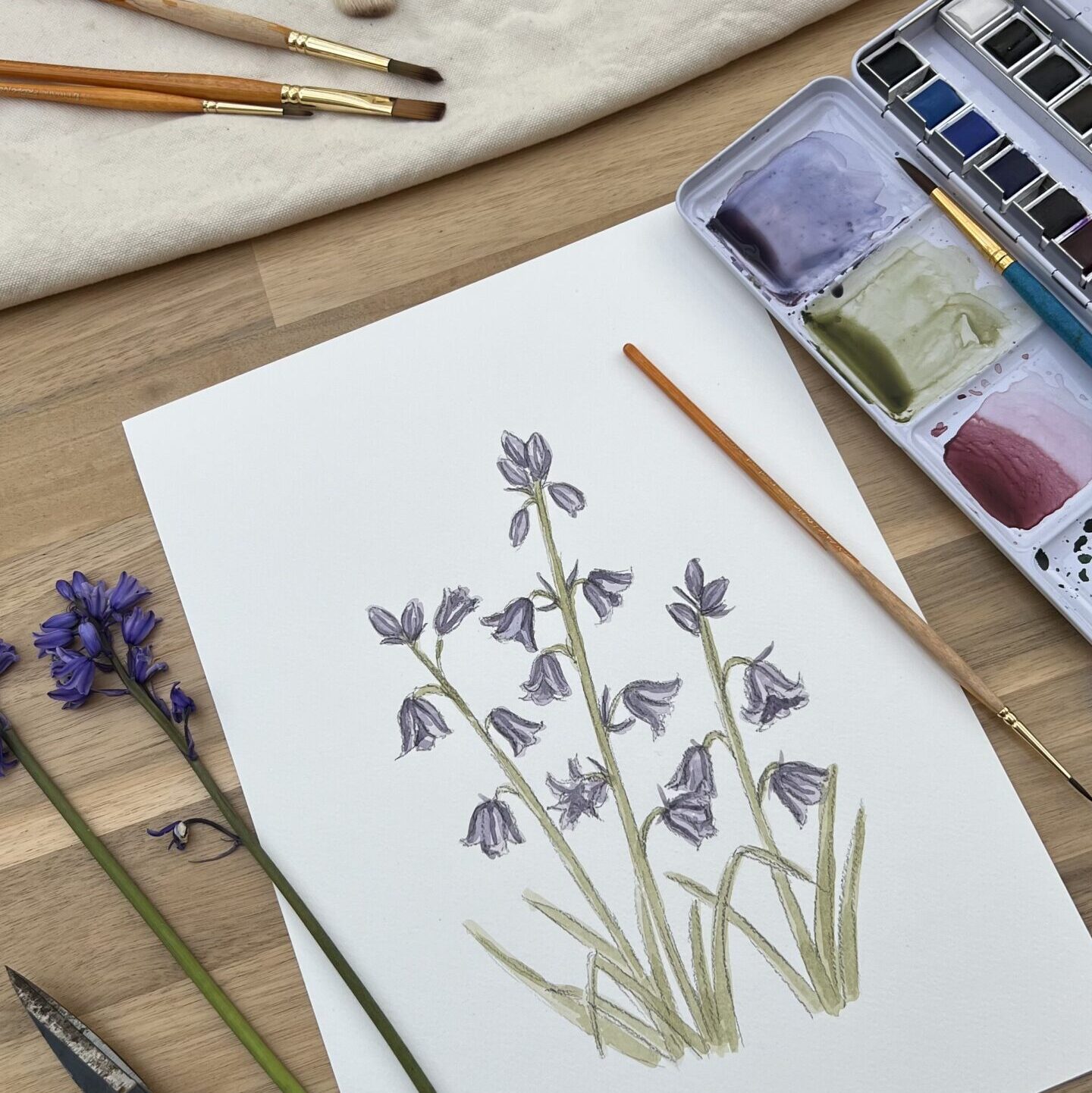 A photograph of a watercolour painting of bluebells from my garden, surrounded by paints and brushes