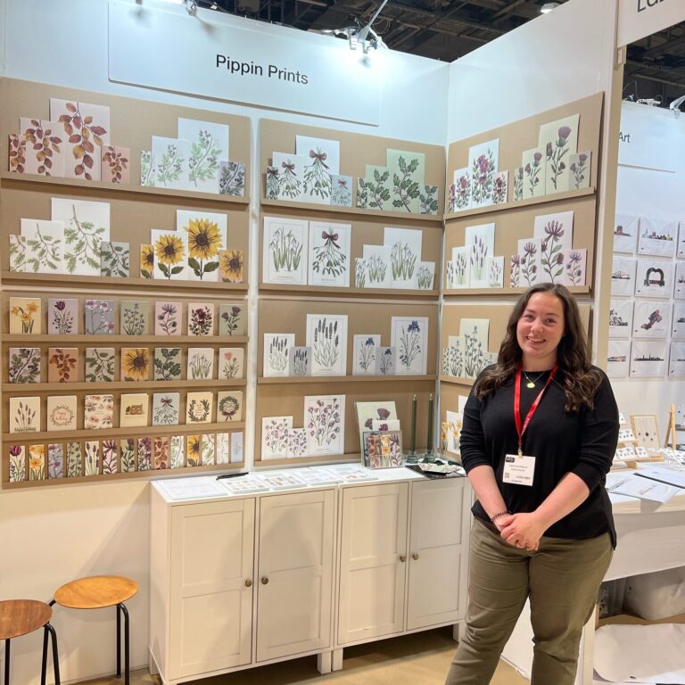 A photo of myself (Emma Macdonald) standing next to my stand at the Autumn Trade Show in Glasgow. My stand is full of my floral cards, prints and bookmarks.