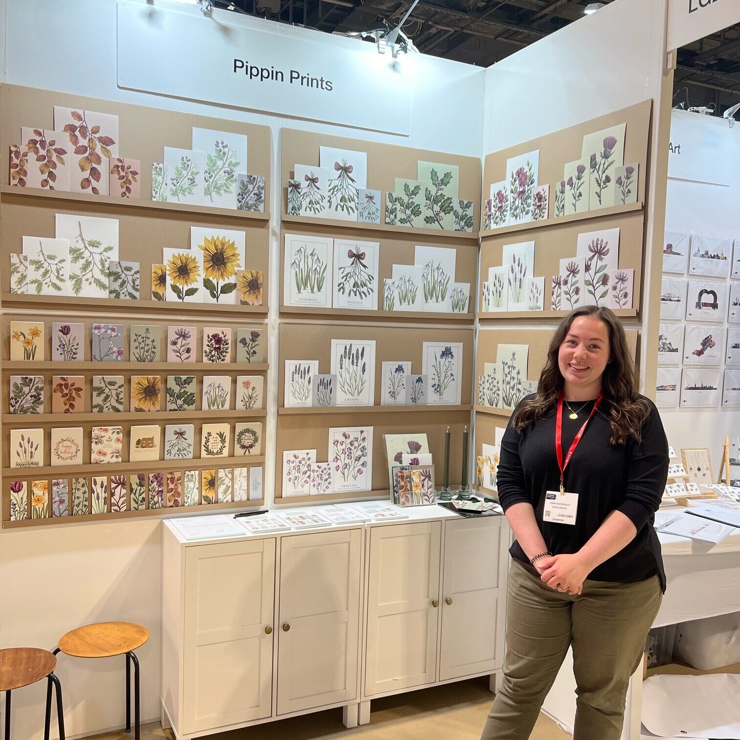 A photo of myself (Emma Macdonald) standing next to my stand at the Autumn Trade Show in Glasgow. My stand is full of my floral cards, prints and bookmarks.